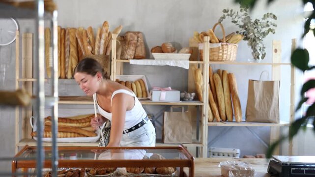 Young female shopper putting fresh croissants in plastic bag in bakery 
