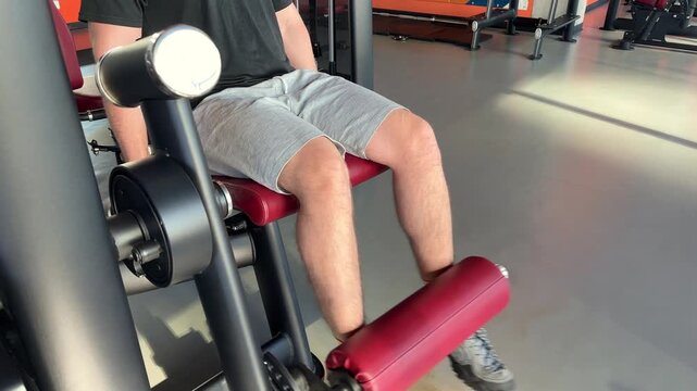 A man performs leg extensions with another adult in the gym background, strength training, lower body workout, active lifestyle.