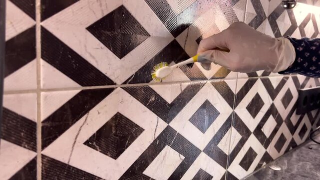 An adult brushes grout between patterned kitchen tiles with a gloved hand, household cleaning, home hygiene, surface care.