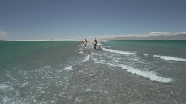 Low altitude head on aerial shot flying forward and over a young couple standing on a pebble spit with crashing waves, flat log profile