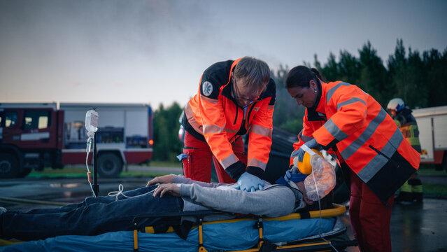 Two Paramedics Perform CPR on an Unconscious Woman Lying on a Stretcher After a Car Crash, One Pressing Her Chest and the Other Giving Oxygen, While Firefighters Work by the Fire Truck in Background.