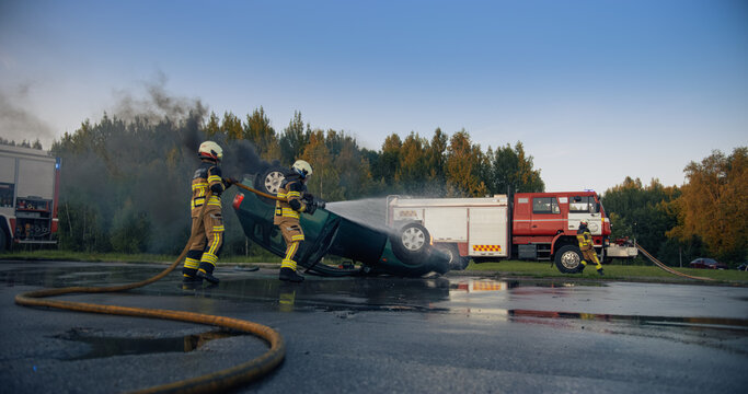 Wide Shot of a Rescue Team Spraying Water on a Flipped Car That Is Smoking After a Road Crash, With a Third Crew Member Running From the Fire Truck to Help on the Wet Highway in the Evening.