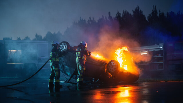 Two Firefighters Use a Hose to Put Out Flames on an Overturned Car After a Traffic Accident on a Road at Night, With a Fire Truck and Emergencys Behind the Crash Site.