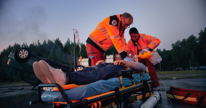 Two Paramedics, a Man and a Woman, Treat an Injured Patient on a Stretcher Beside an Overturned Car After a Traffic Accident, Giving Oxygen and an Iv Drip at the Roadside During an Emergency Call.