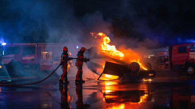 Two Firefighters Aim a Hose at an Overturned Car Engulfed in Fire After a Night Road Crash, Two Fire Trucks Parked on Both Sides of the Street During an Emergency Rescue Call Out.