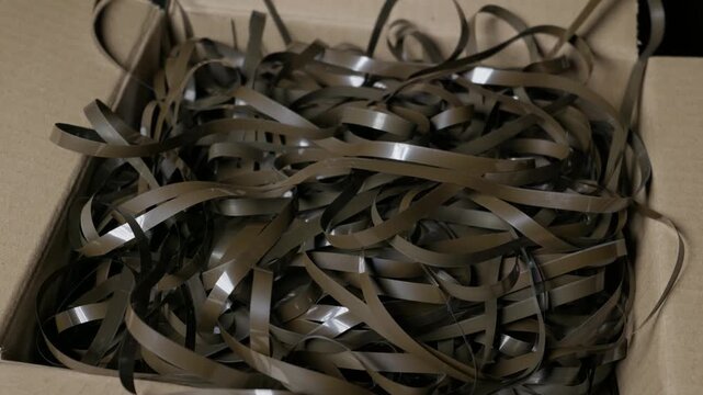 Hands untangle and sort messy magnetic audio tape inside an open cardboard storage box. Soft indoor lighting emphasizes the glossy plastic sheen during the archival restoration process.