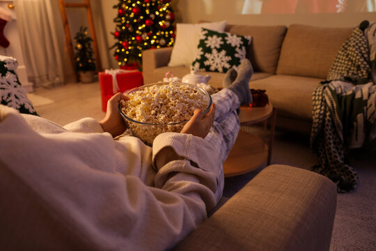 Christmas movie night and holiday relaxation, person in pajamas eating popcorn on sofa at home with festive tree lights and cozy winter atmosphere