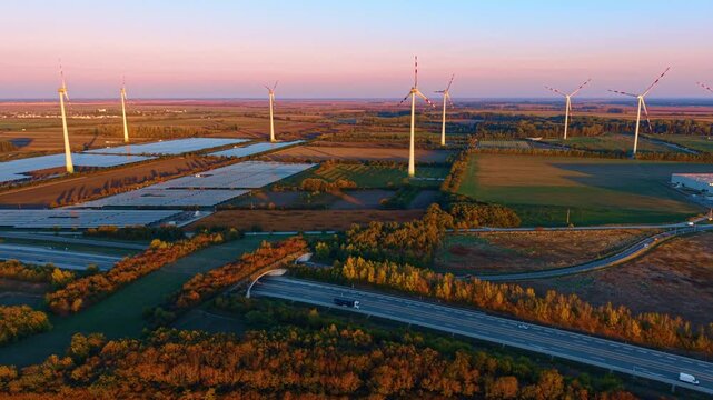 Wind turbines stand above solar arrays beside a highway. Vehicles pass cultivated fields and an overpass beneath a pink band of sky.