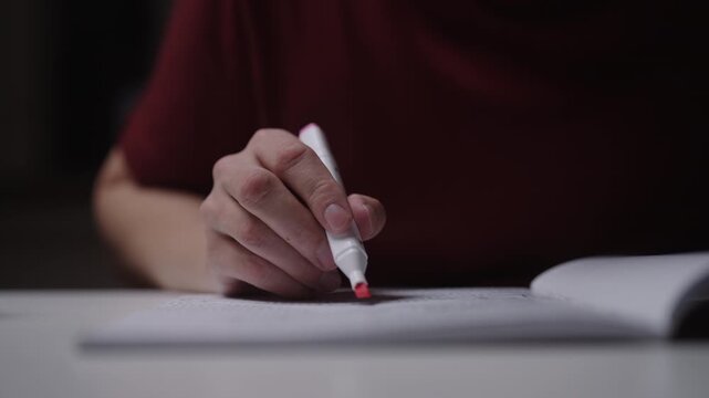 Close-up of student hand opening pink highlighter and underlining text in notebook, studying, preparing for exam sitting at home office table on dark room. Shooting in slow motion.