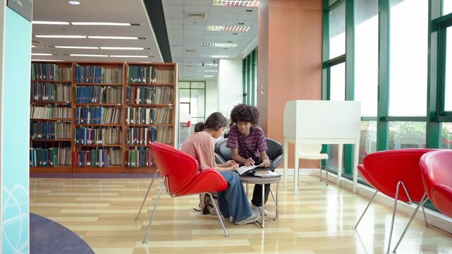 Two young students use tablet while studying at table in modern library. 
