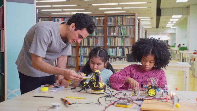 Teacher assists two young girls working on robotics project in library. 