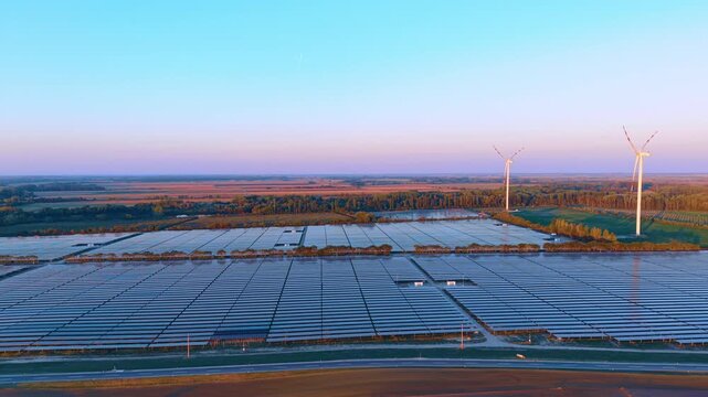 Two wind turbines rise beyond rows of solar panels. A road borders the foreground while access tracks lead toward fields and trees on the right.
