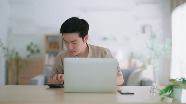 Happy young Asian man working on laptop in living room. Freelance worker male sitting at desk writing notes while watching webinar, studying online
