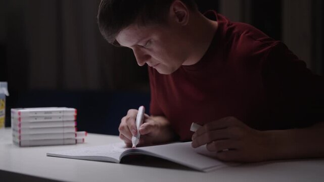 Diligent young artist, student sitting at white table, concentrating on creative hobby, coloring detailed drawing in sketchbook with felt tip pens at night in dark room. Shooting in slow motion.