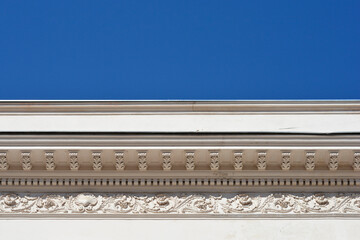 A low angle shot of a white neoclassical building roof cornice featuring a row of decorative corbel brackets and a detailed floral frieze relief under a blue sky. © nahhan