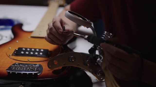 Close-up hands of unrecognizable skilled craftsman soldering potentiometer for electric bass guitar. Luthier repairing instrument electronics in dimly lit, professional workshop, closeup, slow motion.