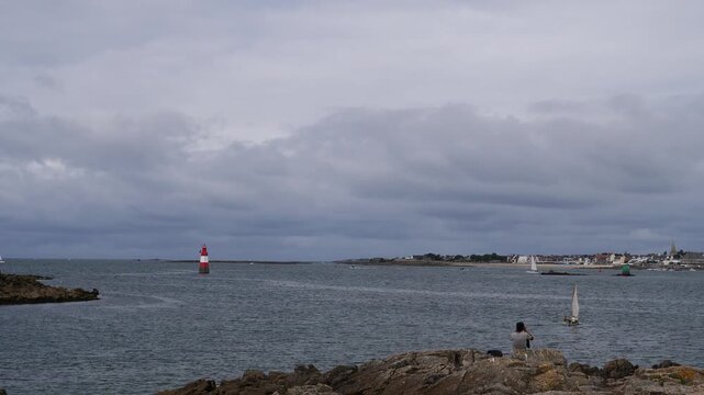 red and white lighthouse turret in calm sea with person on rocky coast under cloudy sky