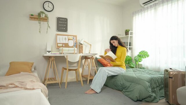 Young Latino female college student sitting on bed and reading a book. 