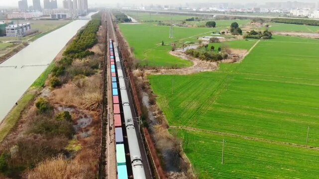 Aerial drone view of a freight train hauling colorful cargo containers along a rural railway line beside a river and green farmland