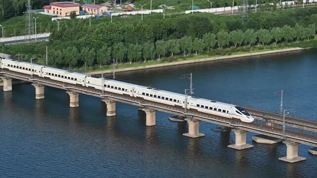 Aerial view of a white high-speed bullet train crossing a multi-pillar concrete bridge over a wide river during daylight