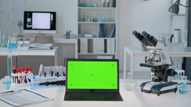 Laptop with green screen mockup standing on research desk beside microscope and chemical test tubes. Perfect for medical software ads, biotech website branding and laboratory technology demos