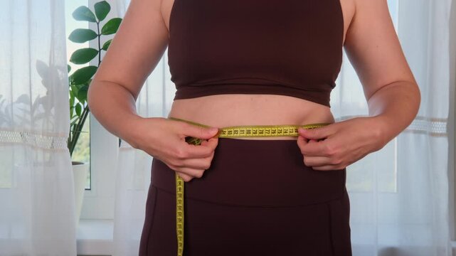 Woman measuring her waist with yellow measuring tape at home. Slim young female in sportswear checking body weight loss results. Fitness diet and healthy lifestyle concept for weight control.