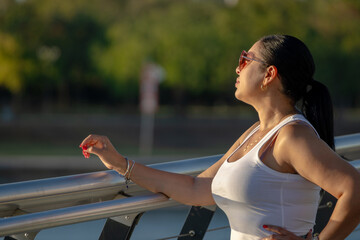 A thoughtful profile portrait of a woman with dark hair tied in a ponytail, wearing stylish sunglasses and a white tank top. She leans against a sleek metal bridge railing during golden hour © Victor