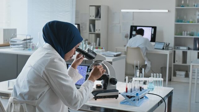 Young adult Asian female scientist examining medical sample with microscope and typing results on laptop in laboratory. Footage ideal for biotechnology and pharmaceutical research campaigns