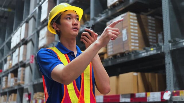Smiling Asian male warehouse worker in safety gear using walkie-talkie and pointing at high shelves. Logistics, inventory control and distribution center concept.