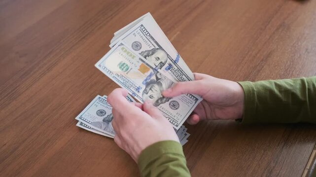 Hands count US dollar cash by sorting hundred dollar bills on wooden table during careful money handling, lifting notes from stack, checking each bill, and organizing cash into neat stack