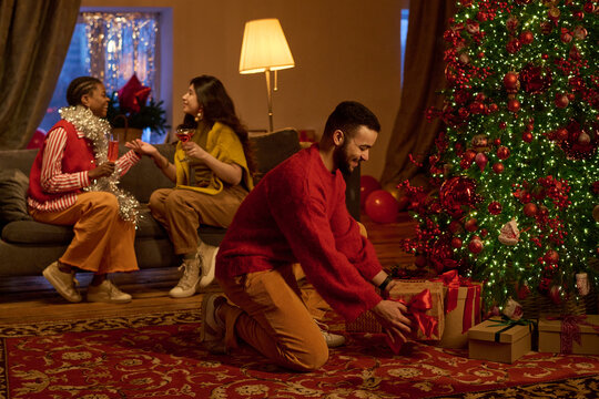 Young adult man placing gift box under decorated Christmas tree near multiethnic friends celebrating winter holidays. Ideal for seasonal advertising and festive retail campaigns