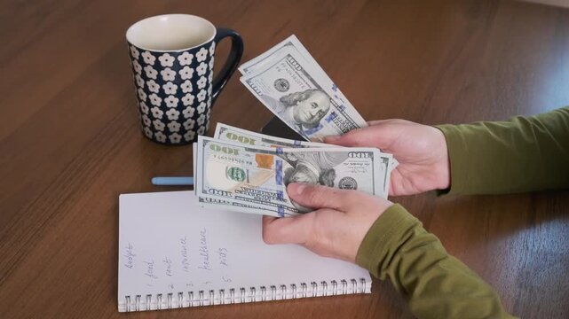 Hands count cash while planning family budget at wooden table, sorting dollar bills above handwritten expense notes and organizing household finances beside patterned coffee mug for monthly spending