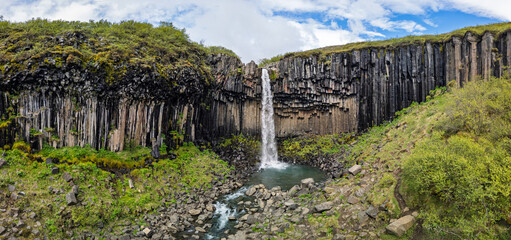 Svartifoss waterfall descends between towering dark basalt columns into a rocky pool, with a winding stream, mossy slopes and open sky at Skaftafell, Iceland, in a summer aerial view. © Artem