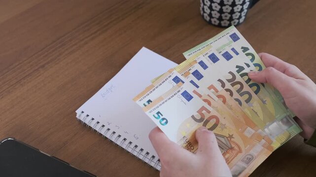 Hands sort and count stack of euro banknotes beside notebook and pen while reviewing family finances, household bills and monthly living costs with limited cash for careful budget planning