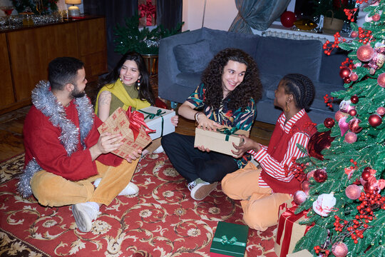 Multiethnic group of young adults exchanging Christmas gifts while sitting near decorated tree. Visual suitable for holiday marketing campaigns and festive retail promotions