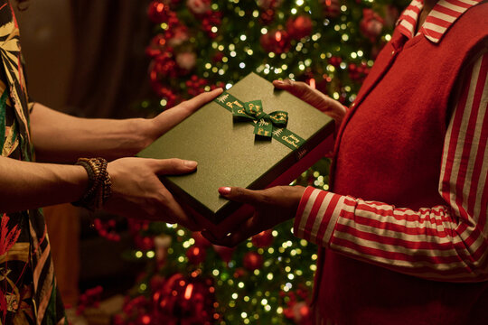Cropped hands of young adult giving wrapped Christmas gift box to Black woman near decorated tree. Useful for holiday shopping campaigns, retail promotions, seasonal festive greetings