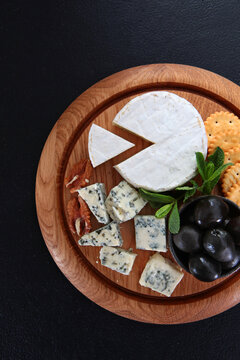 Cheese platter with brie blue cheese crackers black olives walnuts mint on round wooden board. Dark background. Top view