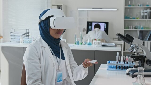 Young adult Asian scientist wearing hijab and VR headset analyzing digital simulation in laboratory. Clip perfect for biotechnology innovations, medical tech ads, and diversity in STEM marketing