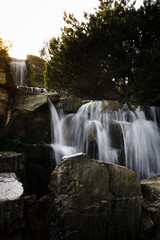 Scenic waterfall cascades over large rocks among lush trees in warm sunlight, creating a tranquil outdoor landscape associated with nature, relaxation and fresh water. © Инна Мадеева