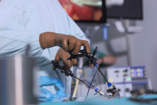 Surgeon hands in brown gloves hold laparoscopic instrument inserting into trocar on patient abdomen sterile blue drapes blurred operating room background clinical procedure