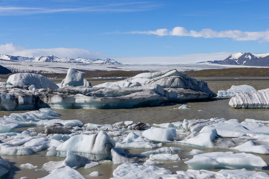 Blue-white icebergs and broken ice float across Fjallsarlon glacier lagoon, with Fjallsjokull and snow-covered mountains beneath a clear blue summer sky in southern Iceland.