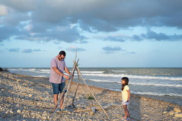 Father arranging natural canes for a teepee shelter while his daughter watches him on a pebble beach at sunset. © Esther Pueyo