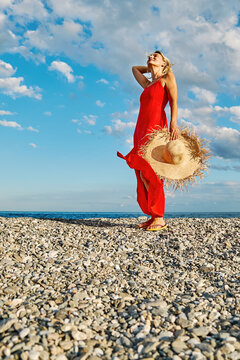 A woman in a red dress holds a straw hat on a pebble beach