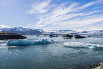 Blue and white icebergs drift across Jokulsarlon glacier lagoon in southeast Iceland, with rippled water, a dark rocky shoreline, distant snow covered mountains and streaked summer sky. © Artem