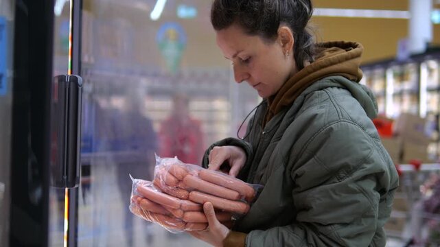 Woman inspects packaged sausages in front of refrigerated display, looking closely at sealed food. Careful grocery shopper considers safe food handling while selecting chilled meat for purchase.