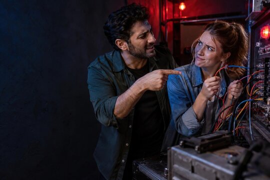 Bearded man pointing at doubtful woman holding multicolored wires at bomb defuse puzzle in escape room, tension