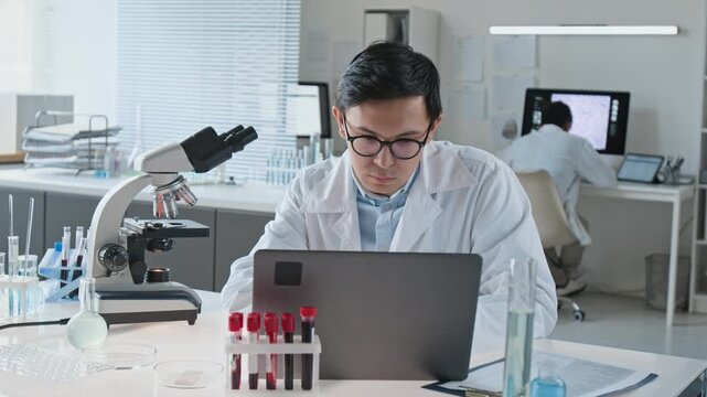 Asian young adult scientist analyzing samples using microscope and typing on laptop in laboratory. Suitable for biotechnology marketing, clinical research videos, and pharmaceutical promo