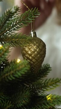 Vertical video. Woman decorating Christmas tree, hanging golden Christmas ornaments.