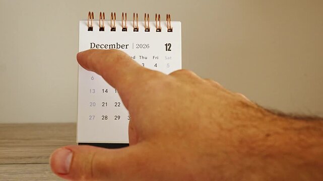 Finger pointing at dates on a white desk calendar showing December 2026, spiral bound pages on a wooden table against a beige wall, concept of year end planning, holiday countdown and final deadlines