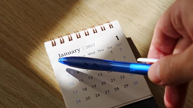 Blue pen pointing at dates on a desk calendar for January 2027, angled top view on a sunlit wooden desk with hard shadows, concept of new year planning, setting goals and organizing the first month